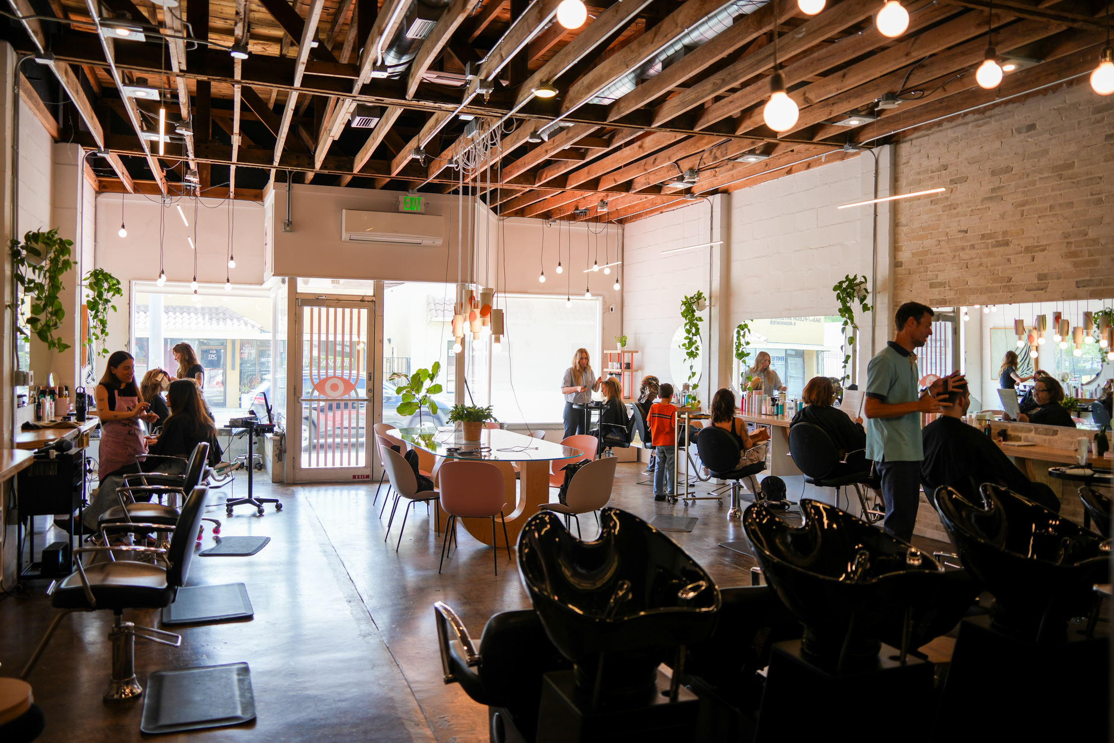 Do or Dye salon interior — wide view of the floor with stylist chairs, washing stations, exposed wood beams, string of bulbs, and the eye-motif entrance